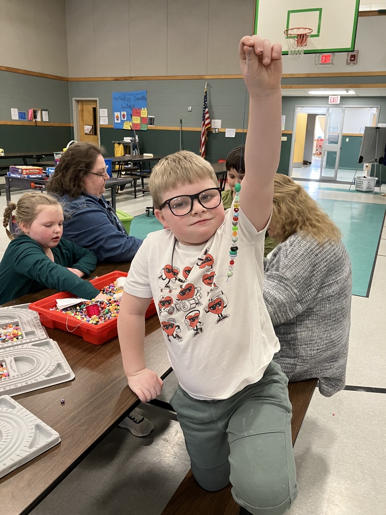 A young boy wearing glasses and a white t-shirt with heart characters stands at a table, proudly holding up a string of colorful beads he has made. Behind him, other children and adults are seated at the same table, which is covered with bead trays and a red bin filled with more beads. The setting appears to be a school gym or community center, with a basketball hoop and an American flag visible in the background.