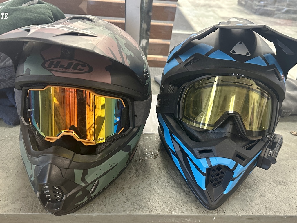 Two full-face snowmobile helmets with goggles rest side by side on a concrete surface indoors.