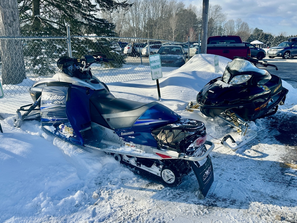 Two snowmobiles parked in a snow-covered school parking lot, surrounded by snowbanks and nearby vehicles.