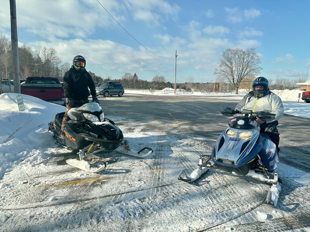 Two students dressed in winter gear sit on snowmobiles in a snowy parking lot, preparing to head out on a clear winter day.