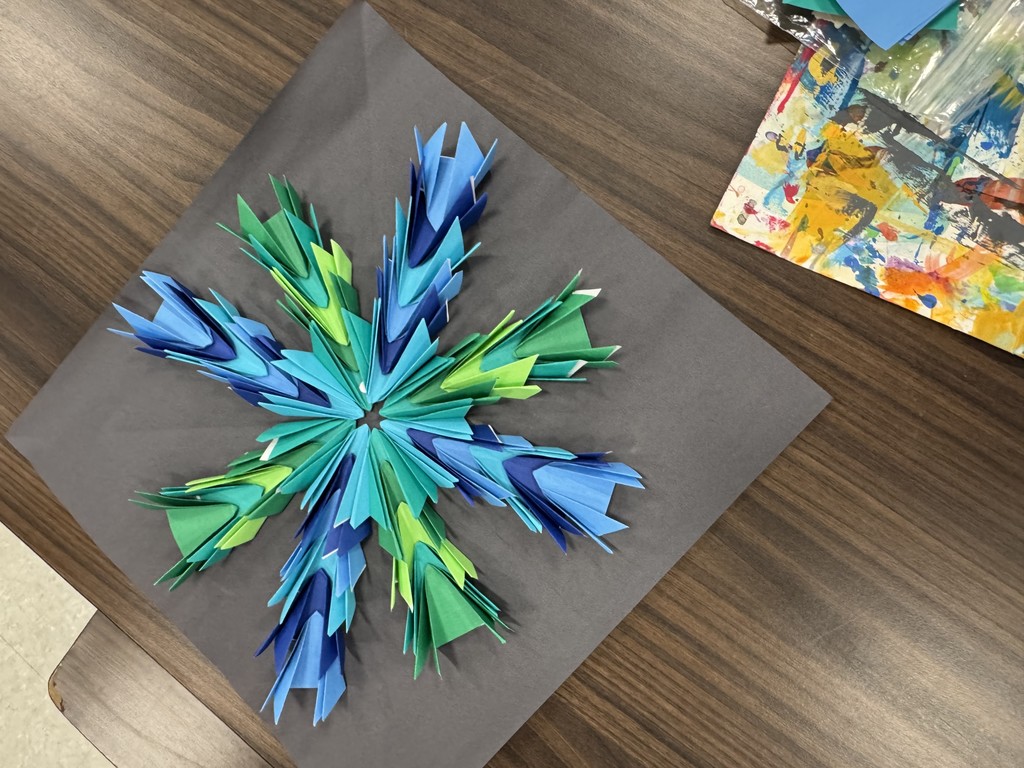 A top-down view of a completed paper-folding project resting on a dark gray square of paper. The artwork is a symmetrical, star-like mandala made of many small, folded "kite" shapes in various shades of blue and green, radiating outward from a central point. The project is placed on a wooden table next to a colorful, paint-splattered sheet and some blue paper scraps.