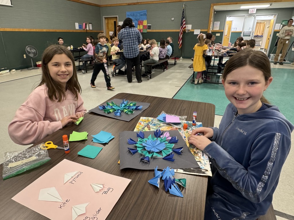 Two young girls smile while sitting at a table in a school cafeteria, working on intricate paper-folding art projects. They are surrounded by colorful paper squares, glue sticks, and completed geometric designs, with a guide sheet on the table labeled "Kite," "Pyramid," "Bug," and "Glider." In the background, other students are seated at long tables eating lunch.