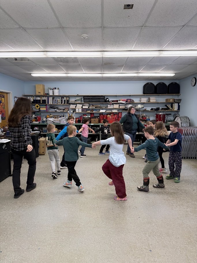 Children hold hands and move in a circle during a movement activity in music class, guided by two adults.