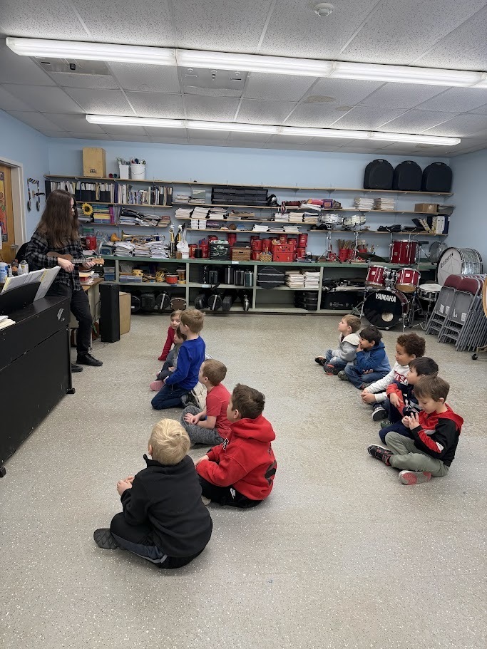 A group of young children sit cross-legged on the floor listening to a teacher holding a ukulele in a music classroom.