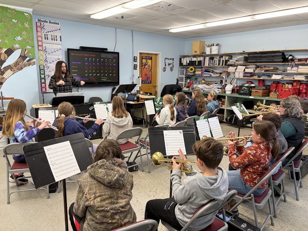 Elementary students sit in rows playing flutes and trumpets during a music class while a teacher conducts from the front of the room.