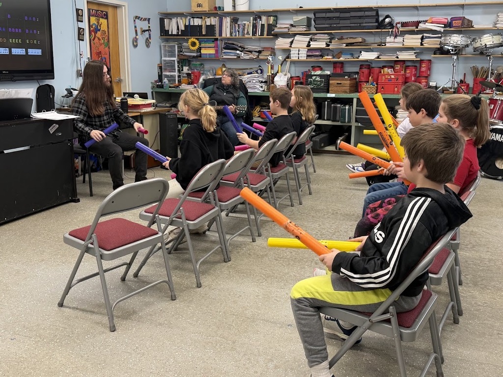 Young students sit in chairs holding colorful rhythm tubes as a teacher demonstrates a percussion activity in a music room.