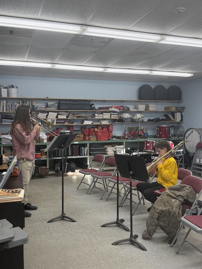 Two students practice trombone facing each other at music stands while a teacher observes in the background.
