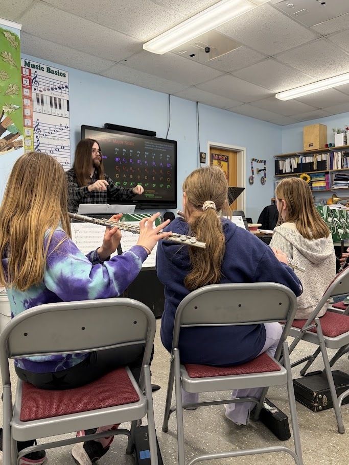 Students play flutes while watching their teacher conduct, with music stands and sheet music visible in the foreground.