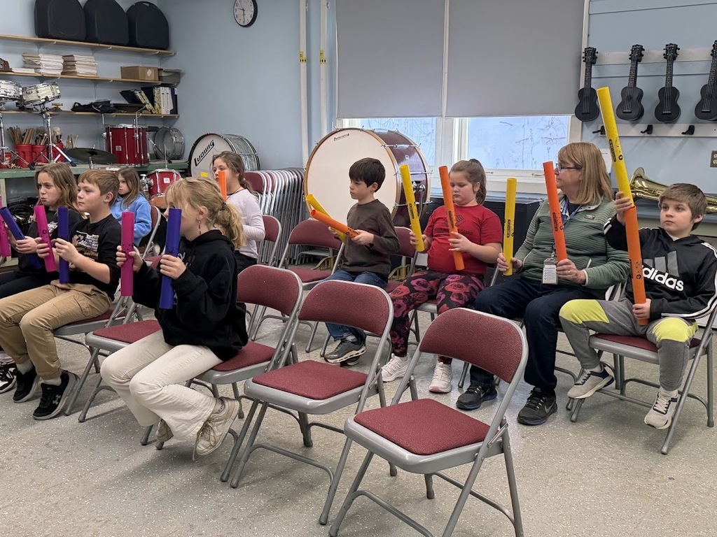 Elementary students hold rhythm tubes upright while following instructions during a group music lesson.
