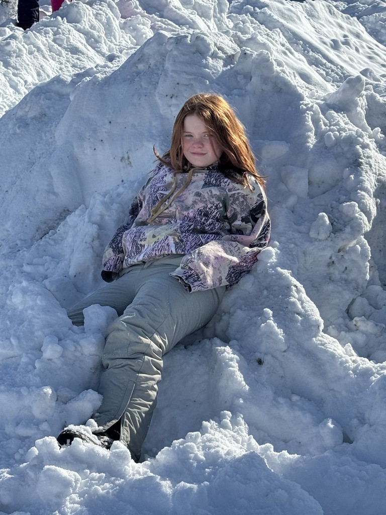 A girl with long reddish hair leans back and smiles while sitting in a large, textured snowbank. She is wearing a patterned camouflage-style hoodie and light-colored snow pants.