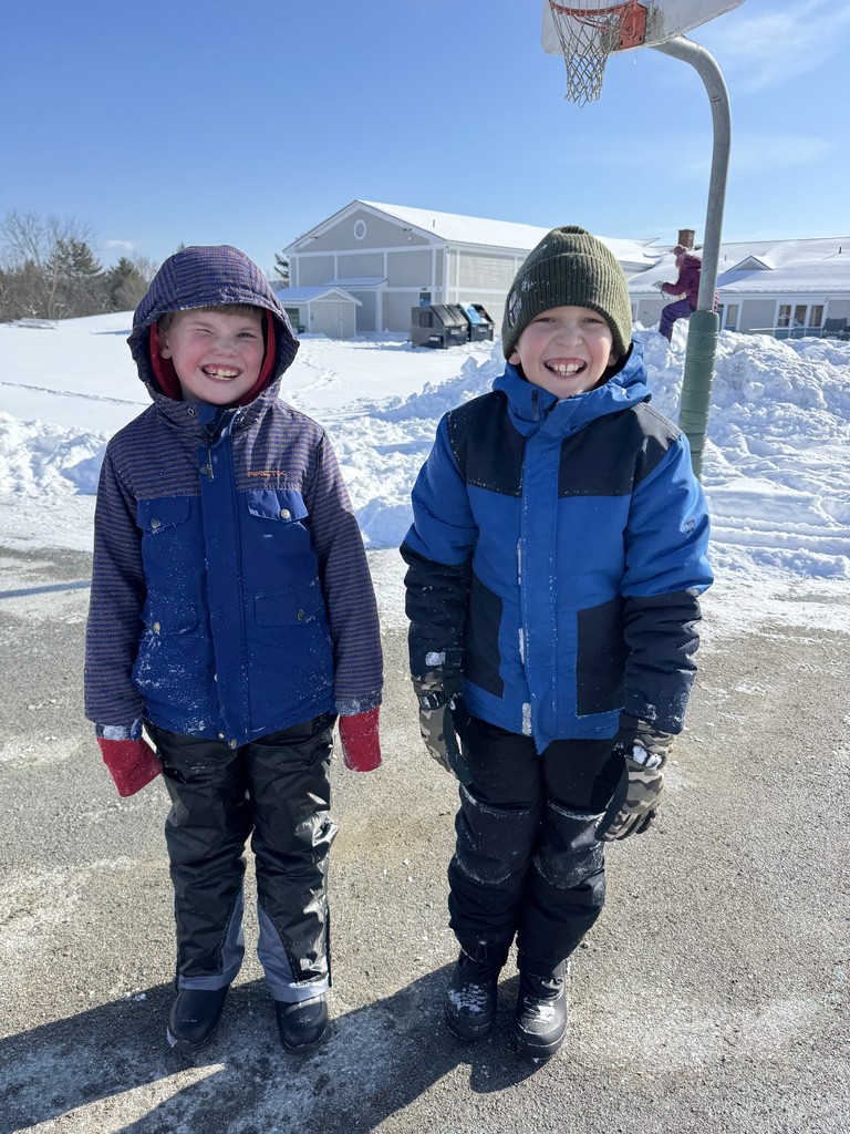Two smiling young boys stand side-by-side on a paved area covered in light snow and ice, wearing heavy winter coats, snow pants, gloves, and hats. Behind them, a snow-covered playground features a large snow pile, a basketball hoop, and a light-colored building under a clear blue sky.