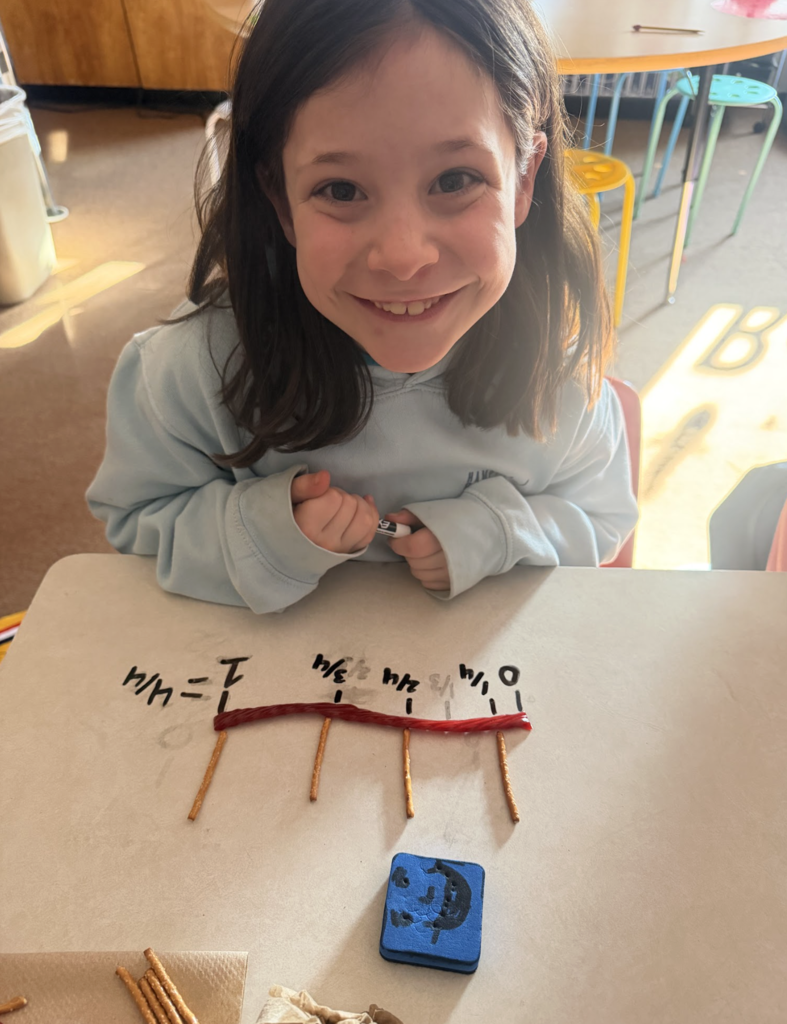 Student smiles at the camera while holding a marker beside a fraction number line made from pretzels and licorice.