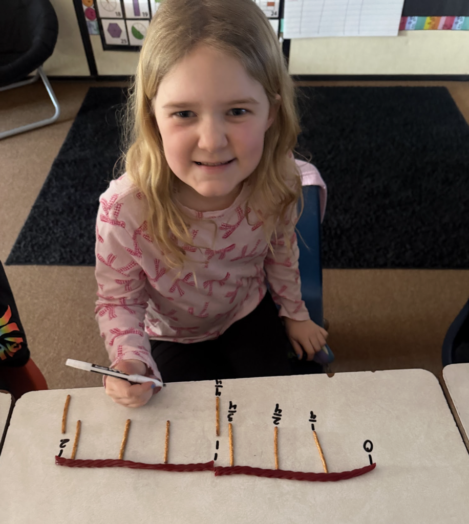 Elementary student smiles while labeling fractions on a number line made with a red licorice string and pretzel sticks.