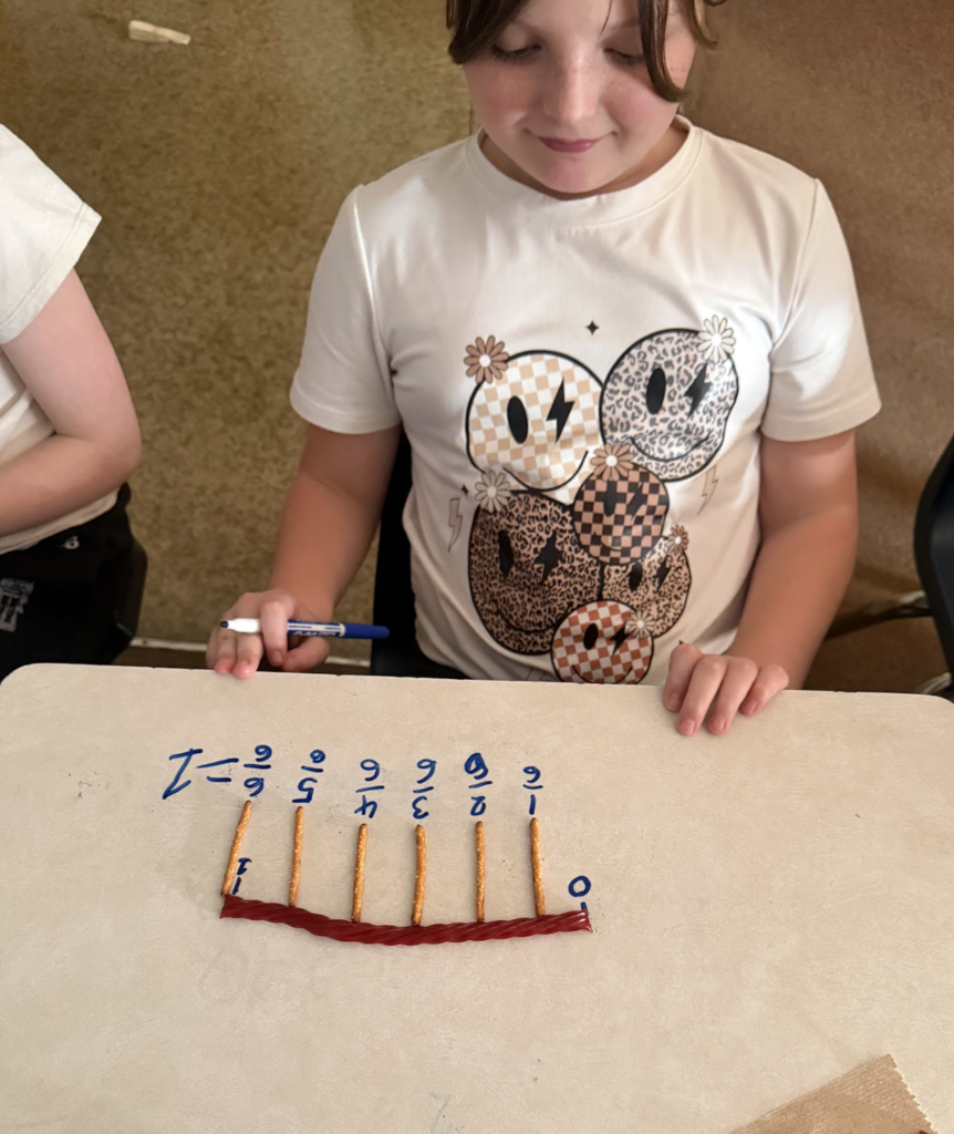 Student holds a marker and looks down at a fraction number line built from pretzels and licorice on a desk.