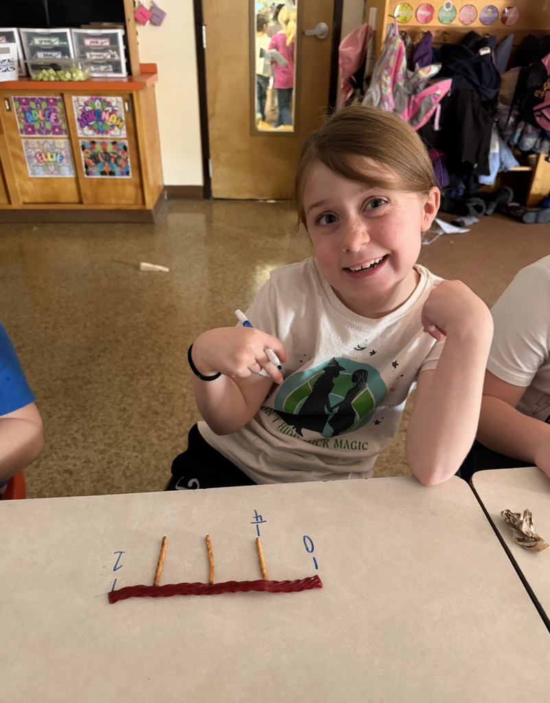 Student smiles and points to herself while sitting at a desk with a pretzel-and-licorice fraction number line.