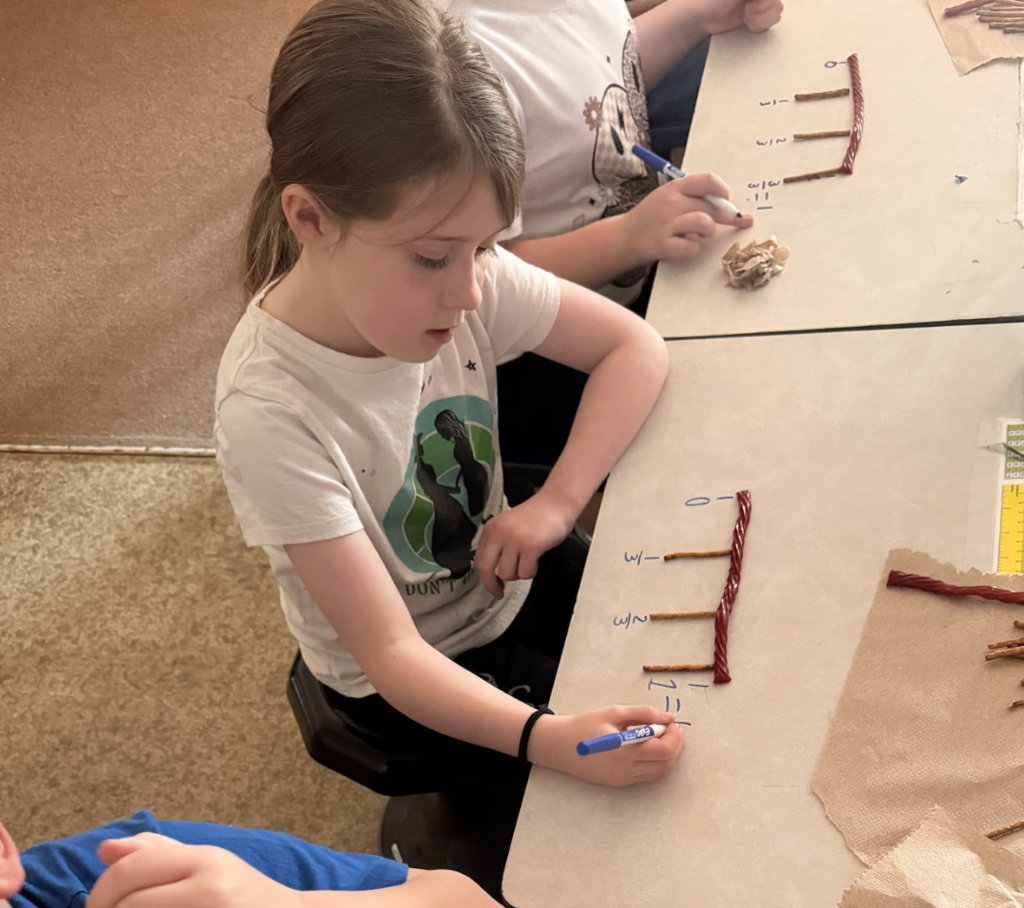 Student smiles while leaning toward a desk displaying a fraction number line made with pretzels and licorice.