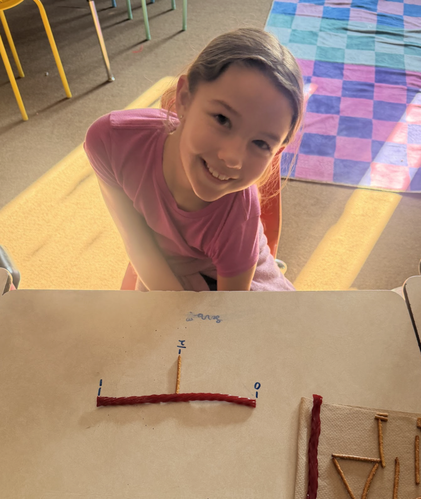 Student smiles while leaning toward a desk displaying a fraction number line made with pretzels and licorice.