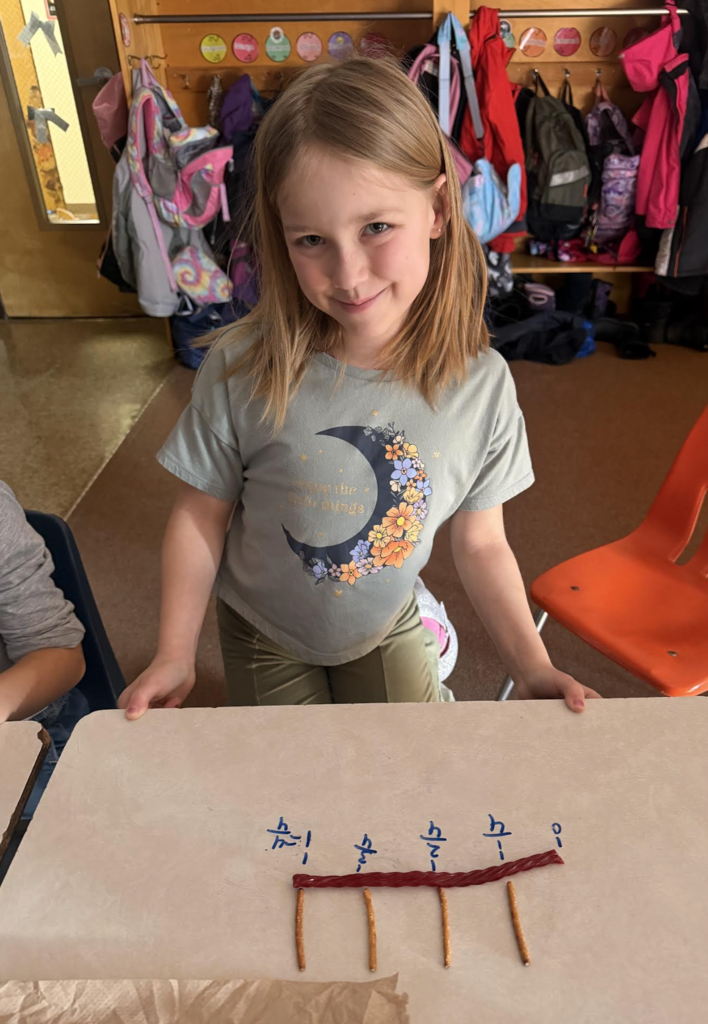 Student stands behind a desk, smiling, with a completed pretzel-and-licorice fraction number line in front of her.