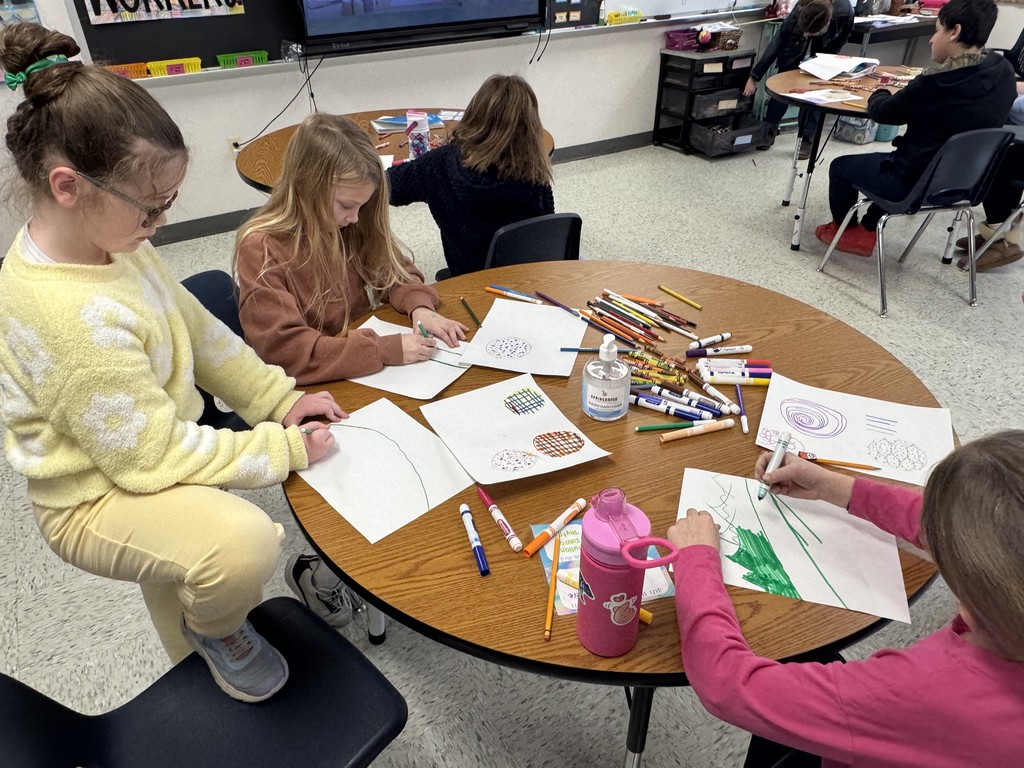 A group of students sits at a large round wooden table covered in markers, colored pencils, and hand sanitizer.