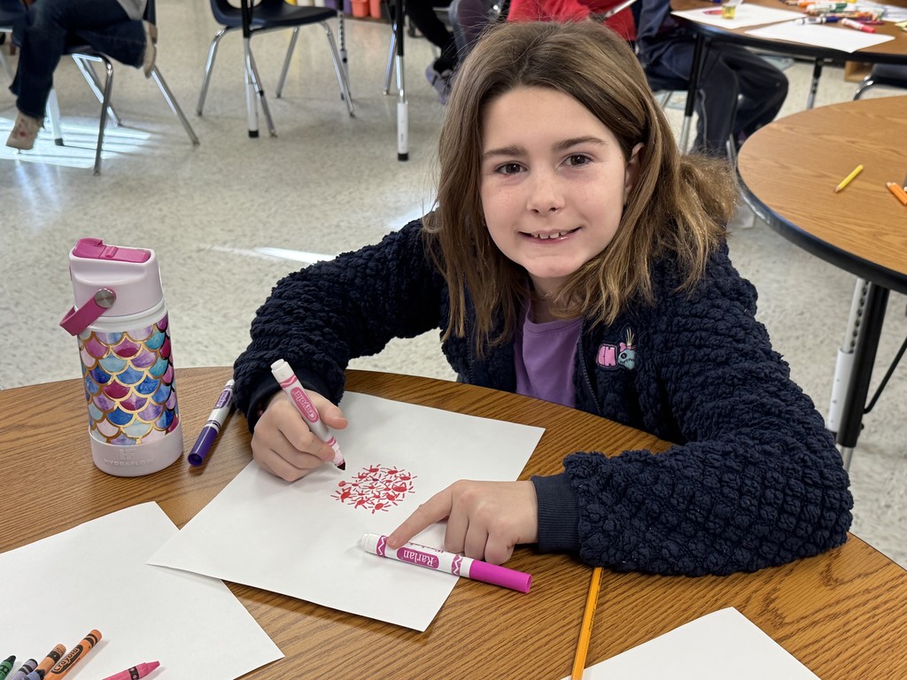 A young girl with brown hair sits at a wooden table in a classroom, smiling at the camera while working on an art project.