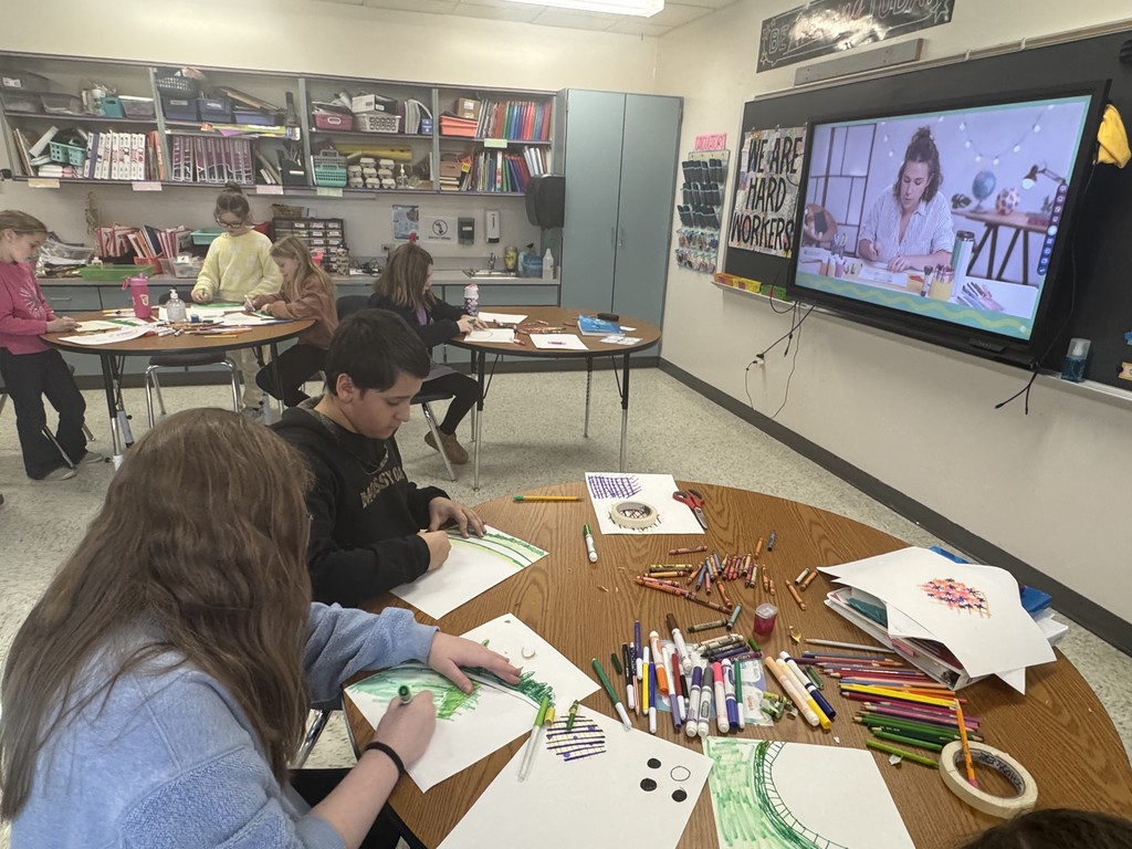 In a bright classroom, several students are seated at round wooden tables, focused on art projects. They are using markers and crayons to draw on white paper, with many art supplies scattered across the tables.