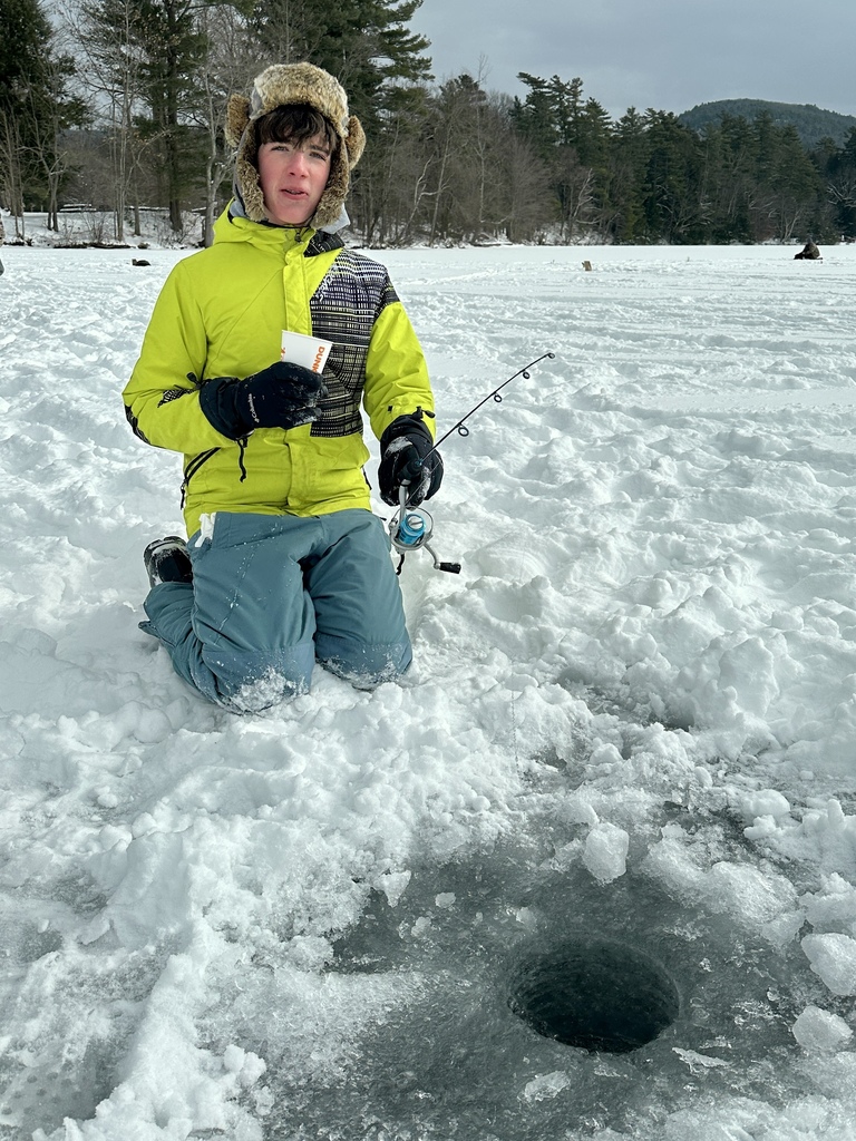 A student in a bright winter jacket kneels on the ice holding a fishing rod and cup, next to a freshly drilled fishing hole.