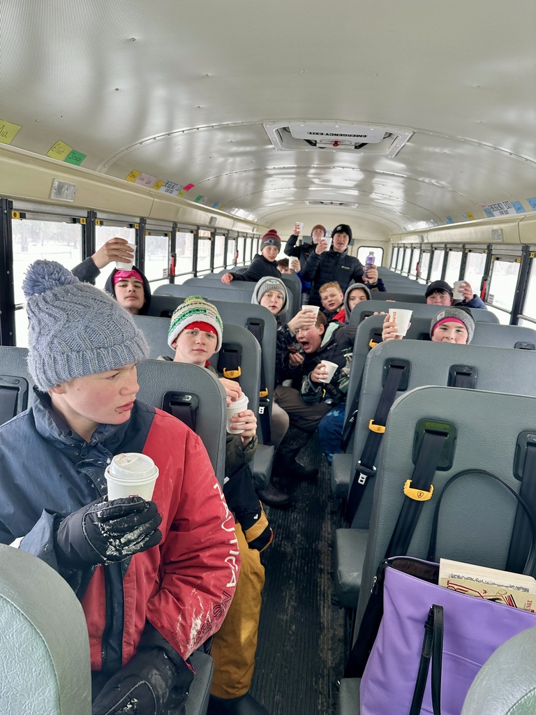 Students seated on a school bus hold up cups of hot chocolate, smiling and bundled in winter clothing after their outdoor activities.