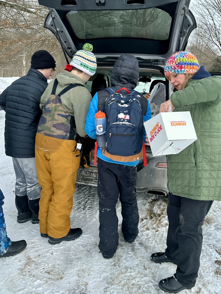 Students gather at the open trunk of a vehicle, reaching for donuts and supplies while dressed in winter gear.