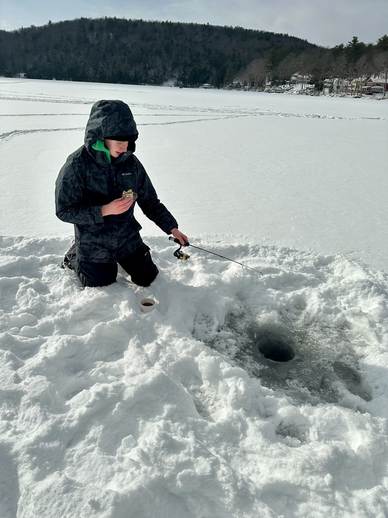 A student kneels on the snow beside an ice fishing hole, holding a fishing rod and snack, with the frozen lake stretching behind them.