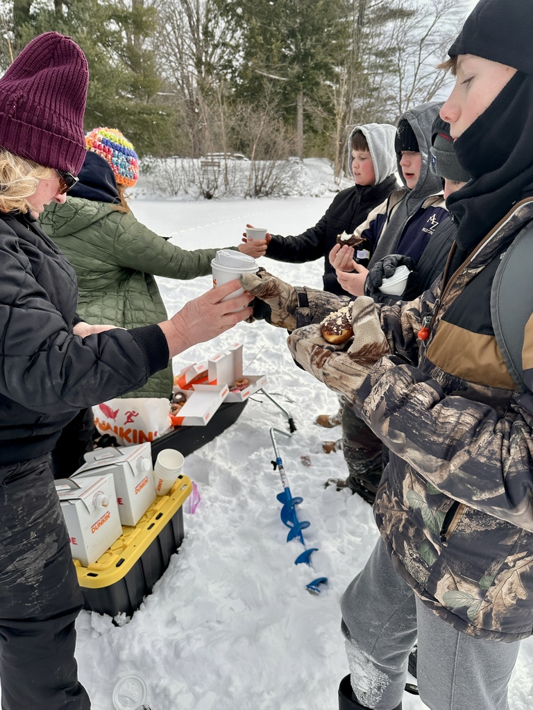 Students wearing hats and gloves accept hot drinks and donuts from an adult while gathered on the snow near ice fishing gear.