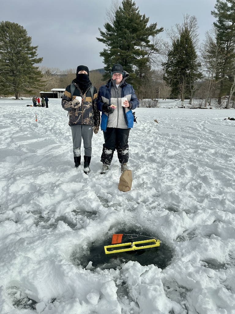 Two students in winter clothing stand on the ice near an ice fishing hole, holding hot drinks and snacks, with snowy trees behind them.