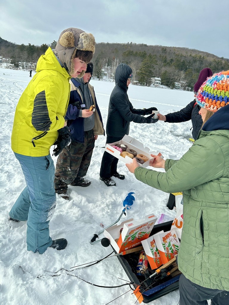 Students and an adult stand on a frozen lake in winter gear as donuts are handed out from an open box beside ice fishing equipment.
