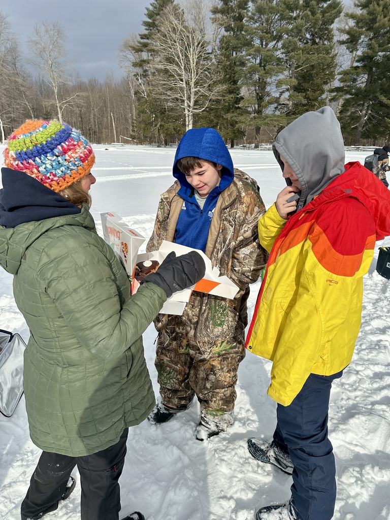 An adult offers donuts from an open box to two students bundled in winter jackets on the snowy lake.