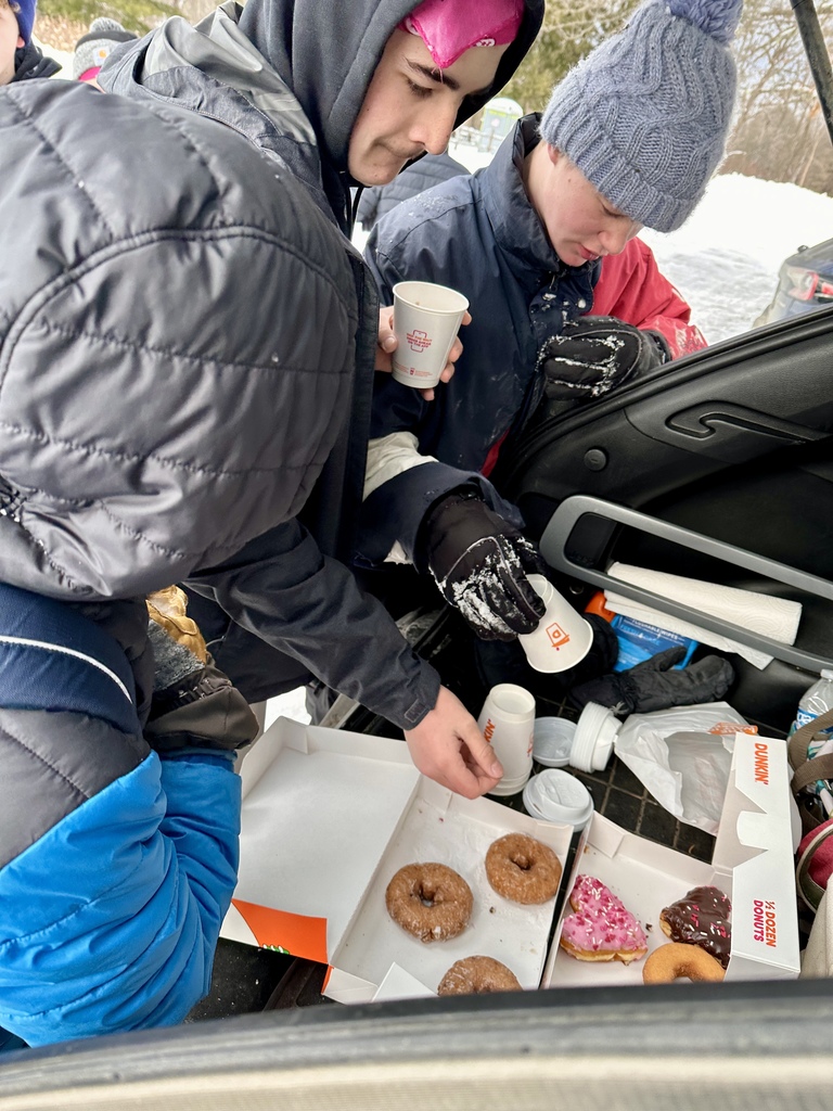 Close-up of students selecting donuts and holding hot drinks at the open back of a vehicle in snowy conditions.