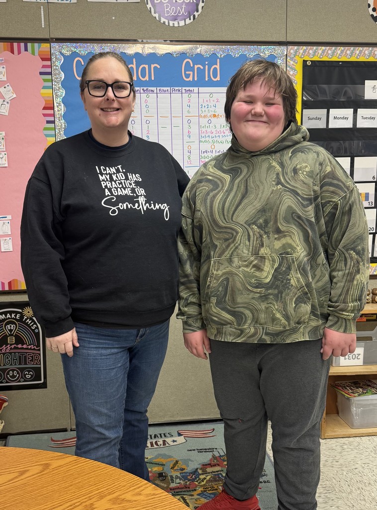 An adult woman and a young boy stand side-by-side in a classroom, smiling at the camera.