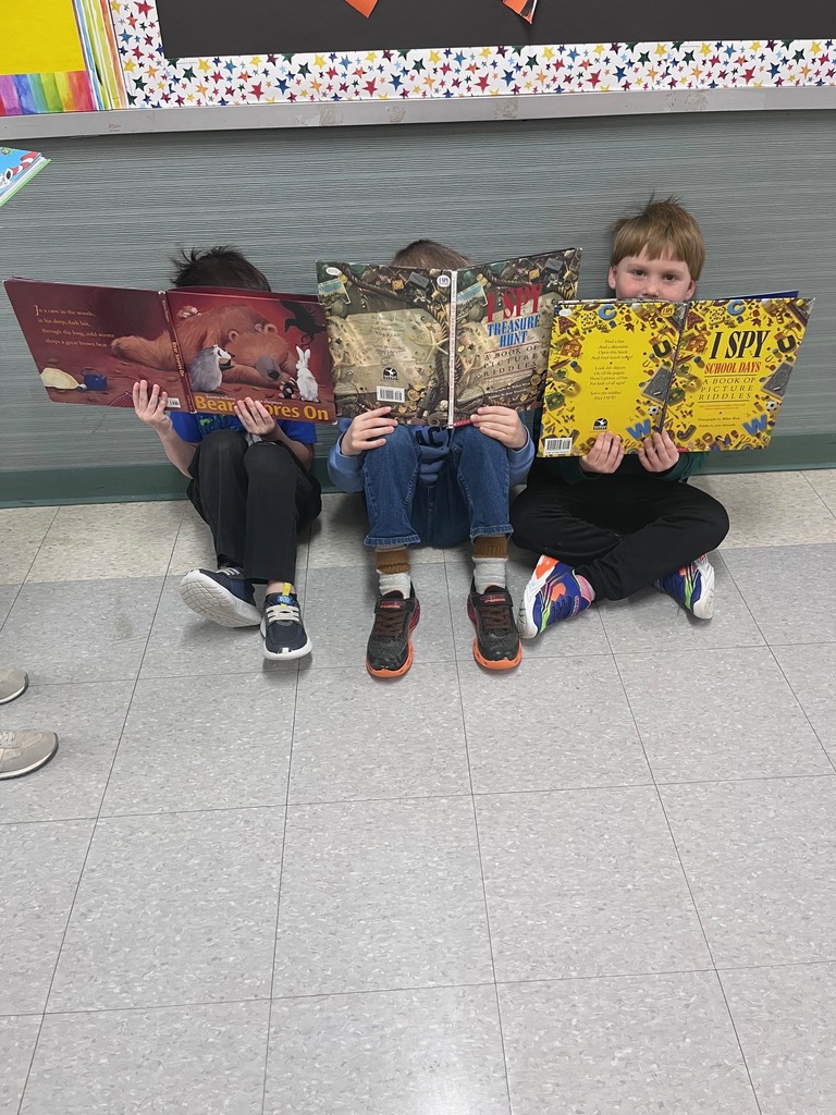 Three young children sit side-by-side on a tiled floor against a gray wall, each holding up a large open children's book