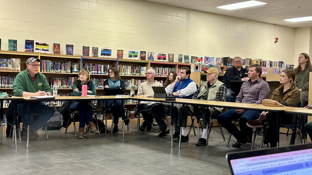 A group of community members sit around tables in a school library, listening to a man speaking at the front, with bookshelves lining the walls behind them.