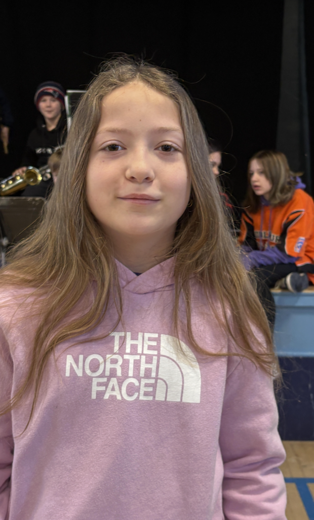 Student with long hair wearing a pink hoodie stands in front of a school stage while other students with band instruments sit in the background.
