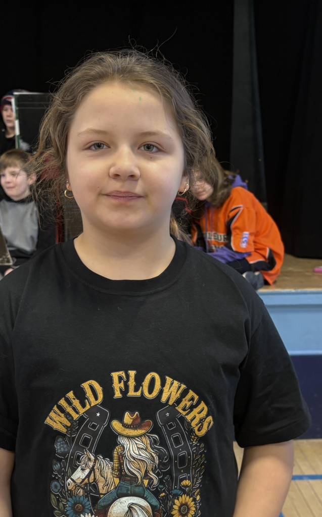 Student wearing a black graphic T-shirt stands facing the camera in a school gym or auditorium with seated students in the background.