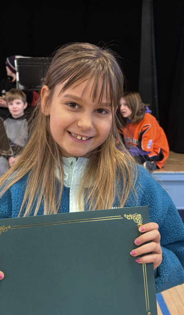 Smiling student in a blue fleece holds a certificate in front of a stage where classmates and music stands are visible behind them.