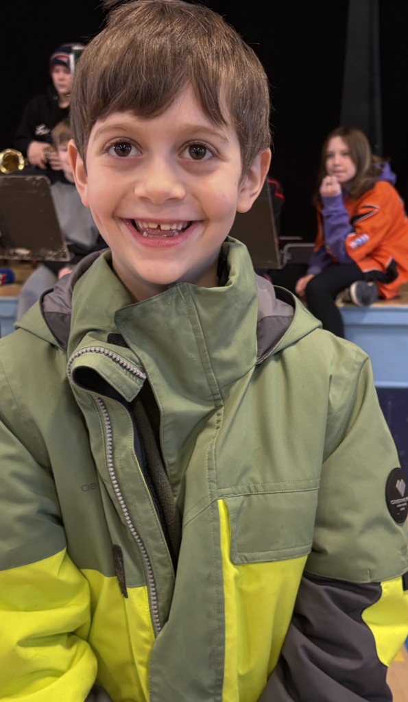 Young student in a green and black winter jacket stands on a gym floor with a school band seated on a stage behind them.