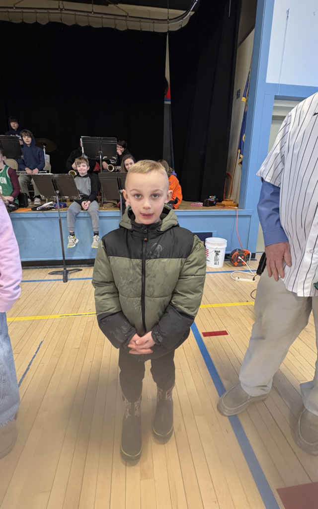 Young student in a green and black winter jacket stands on a gym floor with a school band seated on a stage behind them.