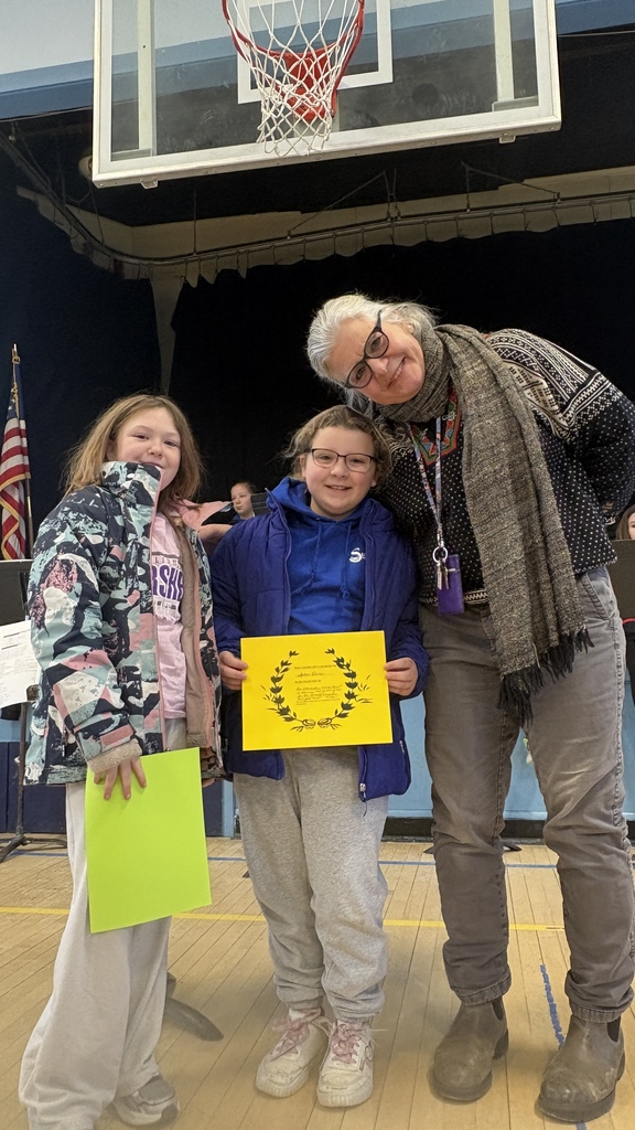Two students and an adult stand together on a gym floor, smiling; one student holds a bright yellow certificate while the other holds a green paper.