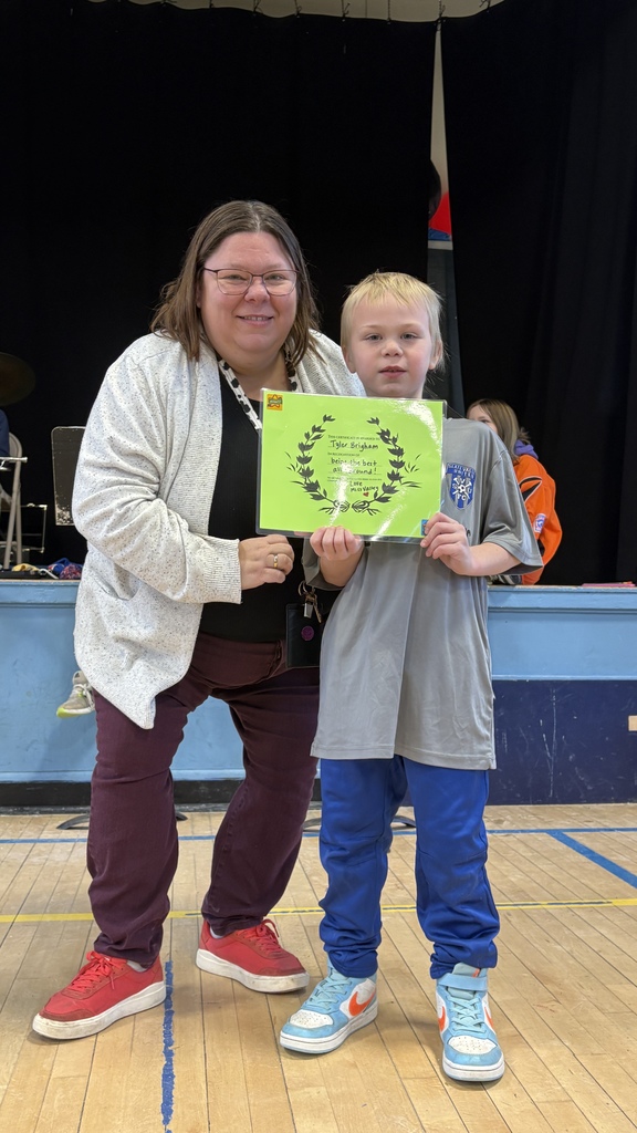Student and adult pose together in a gym with a stage in the background; the student holds a green certificate and smiles.