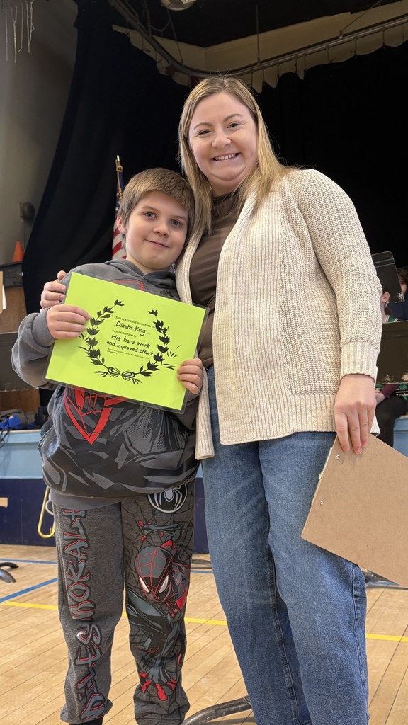 Student and adult stand close together in a gym, smiling; the student holds a green recognition certificate.