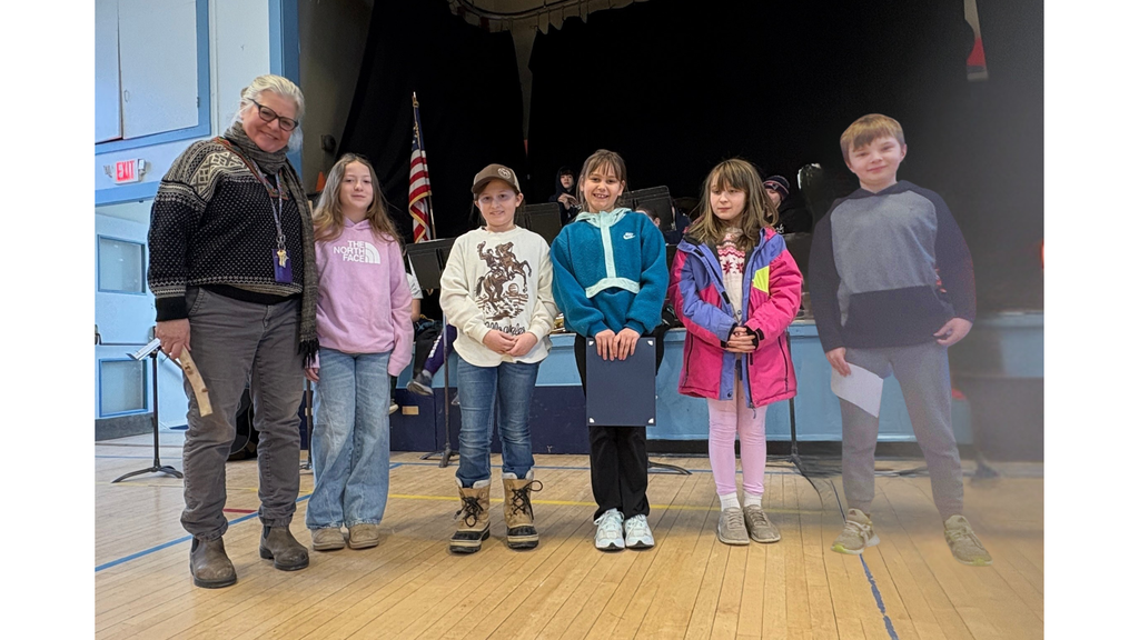Adult and five elementary students stand side by side on a gym floor in front of a stage, smiling for a group photo, with music stands and instruments visible in the background.