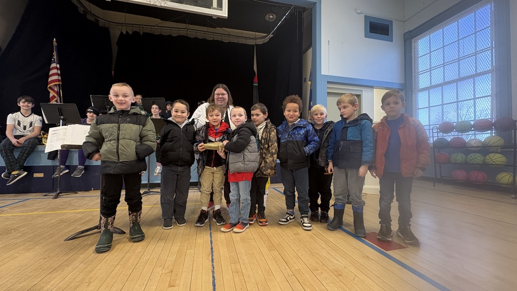 A group of elementary students stand in a line on a school stage with a teacher, smiling after a recognition event, with band students and instruments in the background.