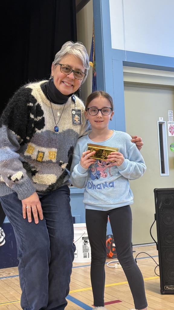 A student holding a gold-wrapped prize stands beside a teacher, both smiling during a school recognition event in the gym.