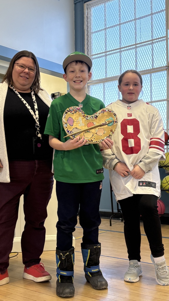 A student in a green jersey holds a decorated art palette while standing between a staff member and another student in a sports jersey, posing in a school gym.