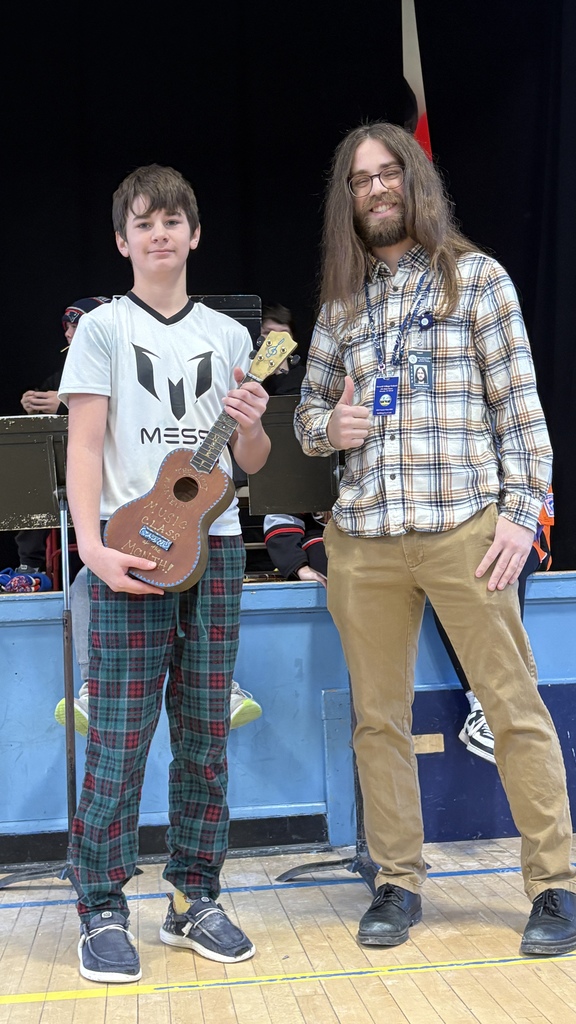 A student holds a small ukulele next to a smiling teacher giving a thumbs-up, with band equipment visible on the stage behind them.